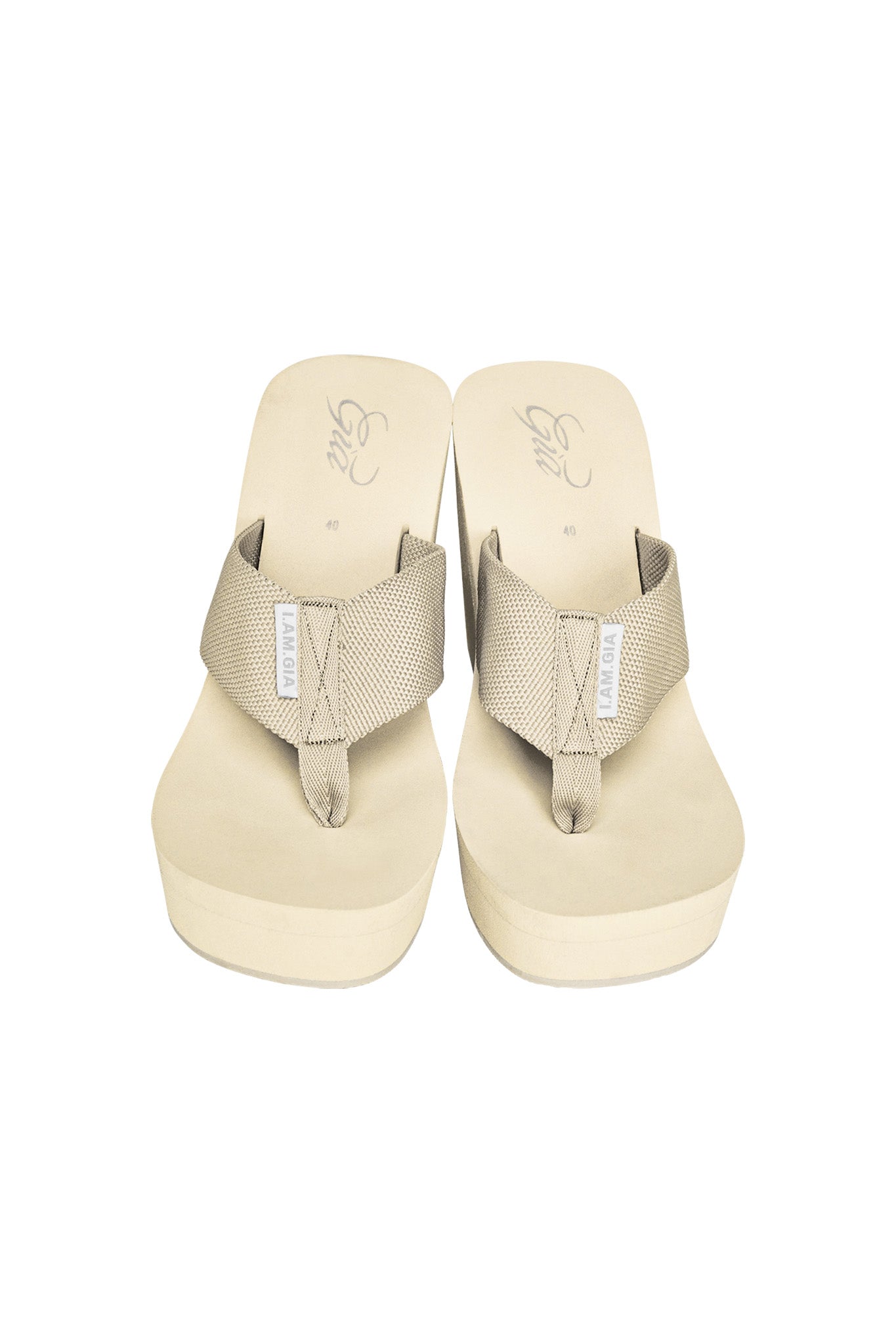 A pair of tan Shelby flip-flops from I.AM.GIA with thick fabric straps and slightly elevated soles. The brand name "I.AM.GIA" is visible on a small tag on each strap. The neutral-toned sandals are placed side by side, showcasing their minimalistic design against a white background.