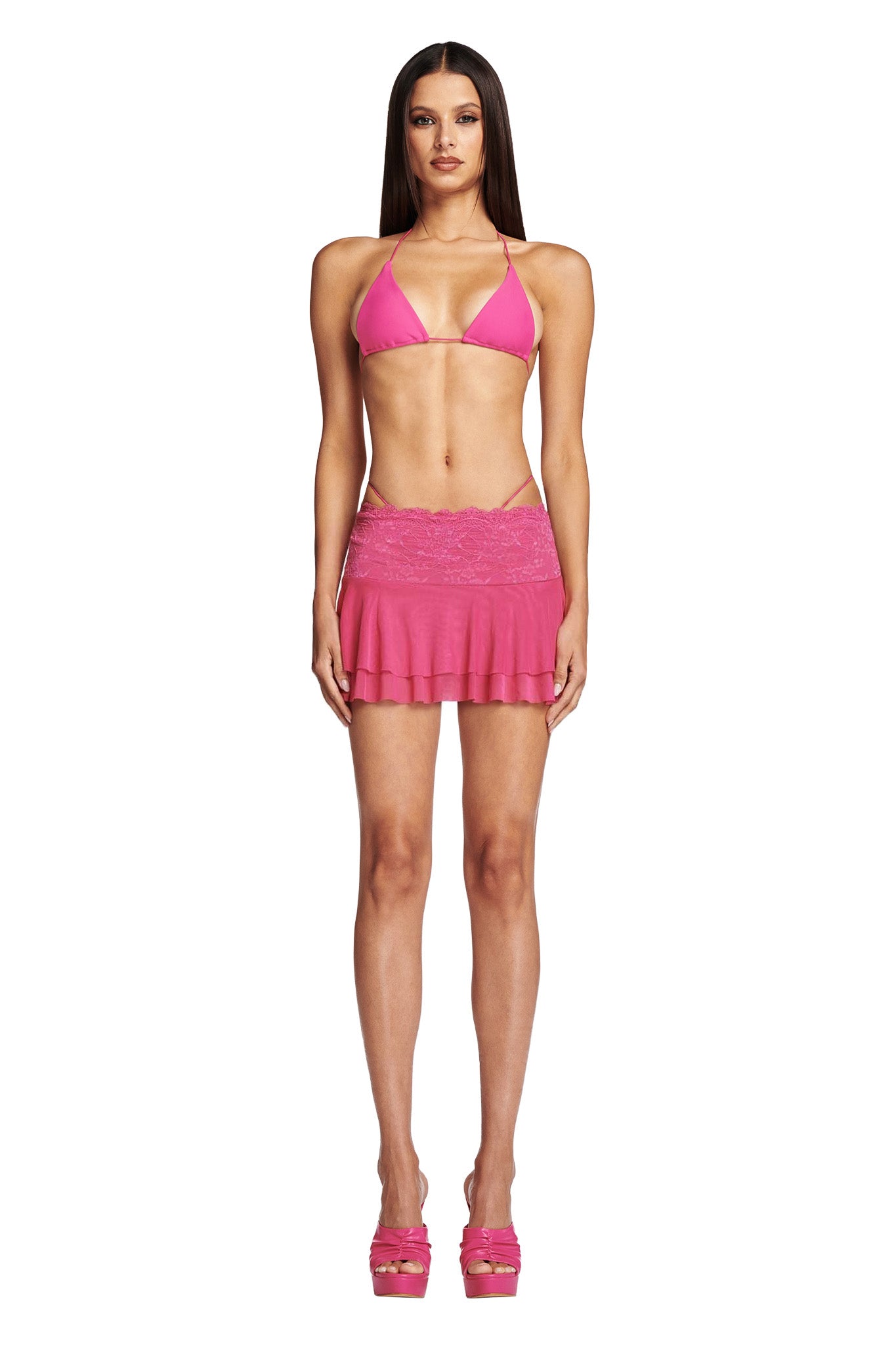 A woman stands wearing a bright pink bikini top paired with the I.AM.GIA BAMBI BIKINI BOTTOM in hot pink and a matching mini skirt. She complements her look with high-heeled pink sandals. The white background makes her outfit stand out prominently. With long, straight hair and a relaxed expression, she exudes effortless style.