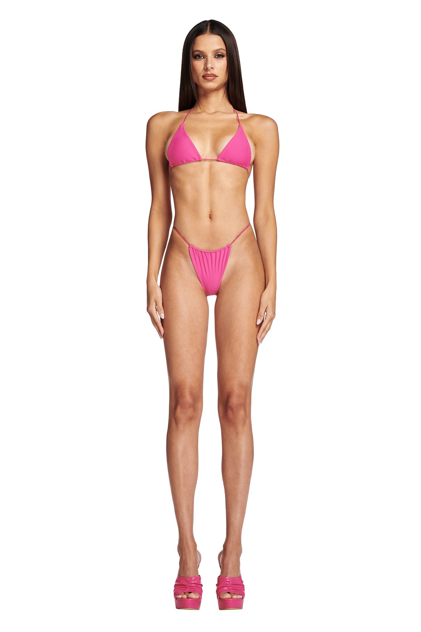 A woman is standing facing forward, wearing the I.AM.GIA BAMBI BIKINI BOTTOM in hot pink with matching platform heels. She has long, straight hair and a neutral expression. The background is plain white.