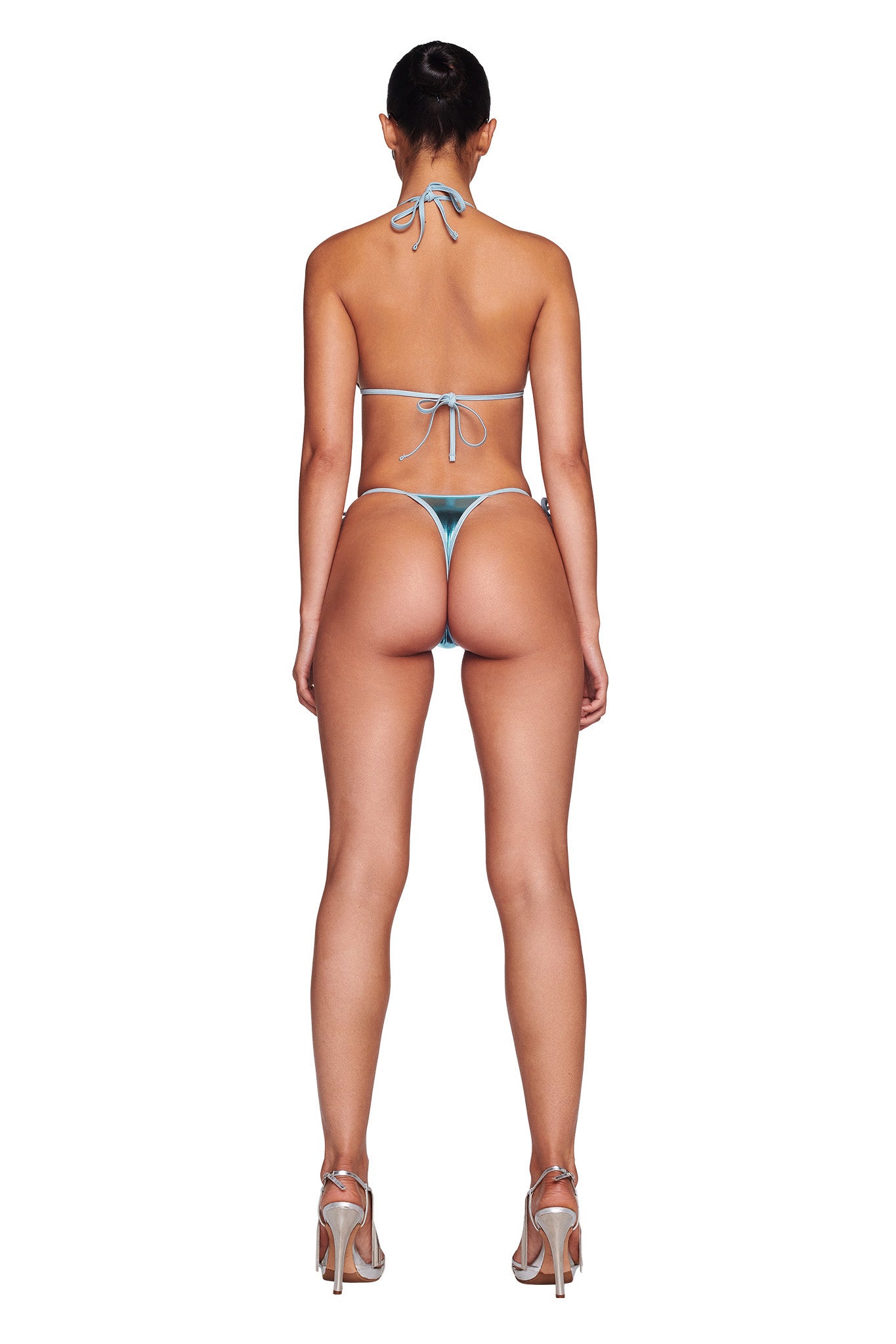 A person is standing against a white background, seen from the back, wearing a light blue ALOHI BIKINI TOP by I.AM.GIA and high heels with their hair tied up. The bikini top features thin straps tied around the neck and back, revealing much of the person's lower back and legs.