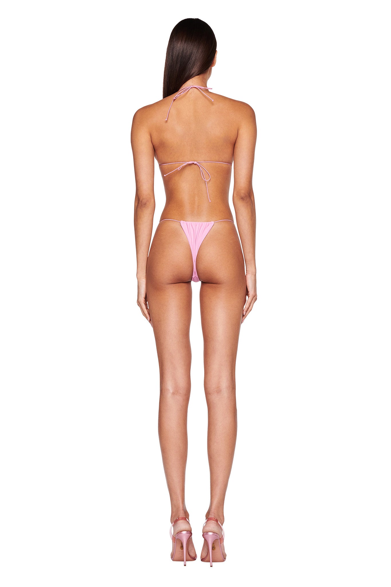 A person with long dark hair is standing with their back to the camera, wearing an I.AM.GIA BAMBI BIKINI TOP in Bubblegum Pink and matching pink high-heeled shoes. The bikini features a halter neck strap. They are standing against a plain white background.