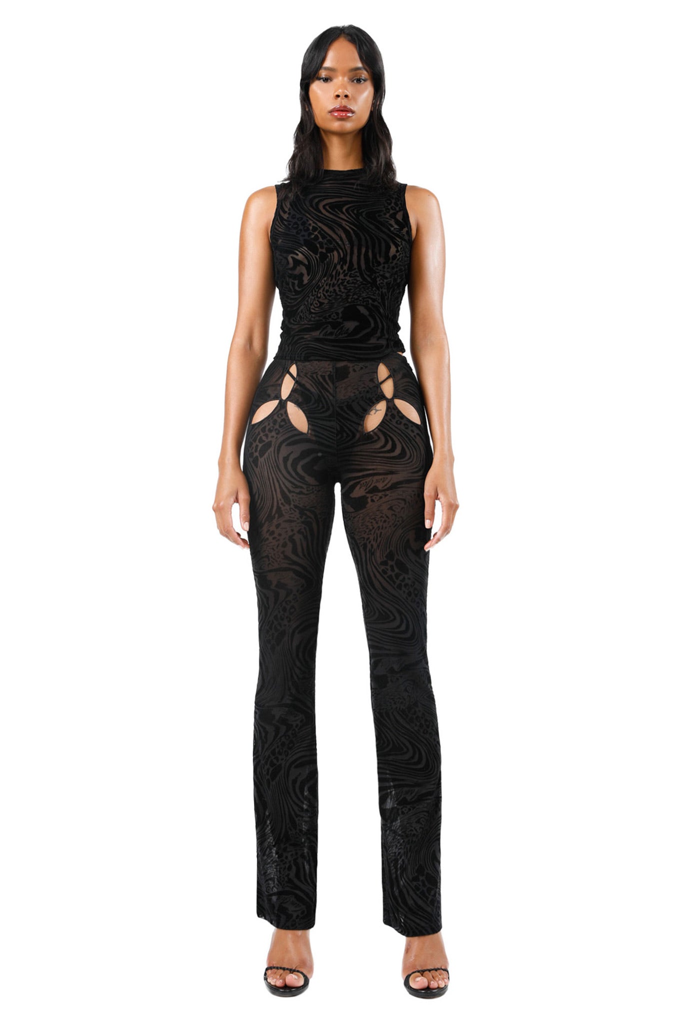 A person is standing against a white background, wearing the AMINA PANT - BLACK BURNOUT by I.AM.GIA. The pants feature intricate patterns with multiple cutouts around the hips and are paired with a high-neckline top. They have long, dark hair and are wearing black open-toe heels.