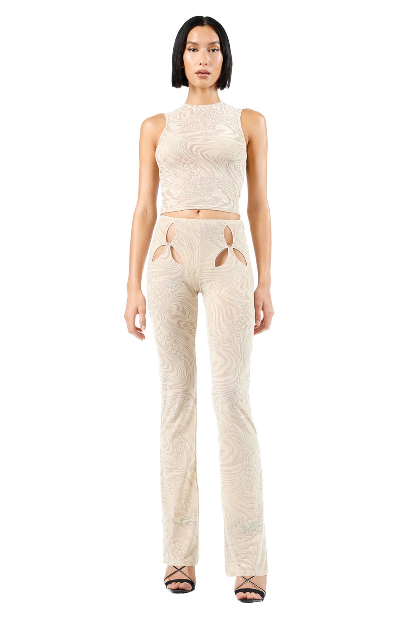A person with straight black hair in a middle part is wearing an I.AM.GIA sleeveless, cream-colored crop top and matching AMINA PANT - TAN BURNOUT high-waisted, flared pants with leaf-shaped cut-out details near the hips. They are standing against a white background and wearing black strappy high heels.