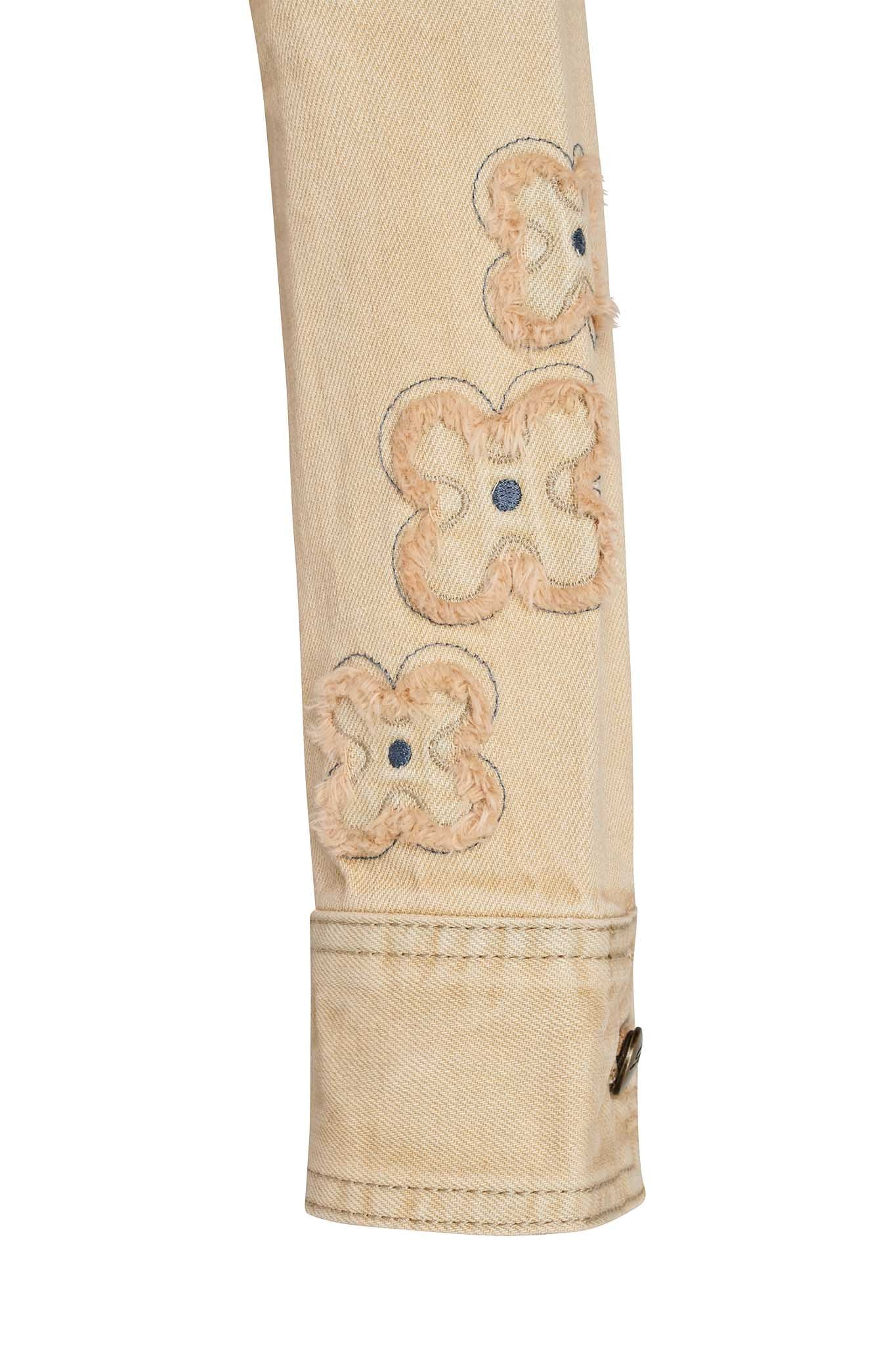 Close-up of the sleeve of the FAUNA JACKET - BEIGE : TAN DENIM by I.AM.GIA, featuring frayed floral patches sewn along the side. The sleeve has a cuff with a button. The visible portion of the jacket highlights its textured and distressed details.