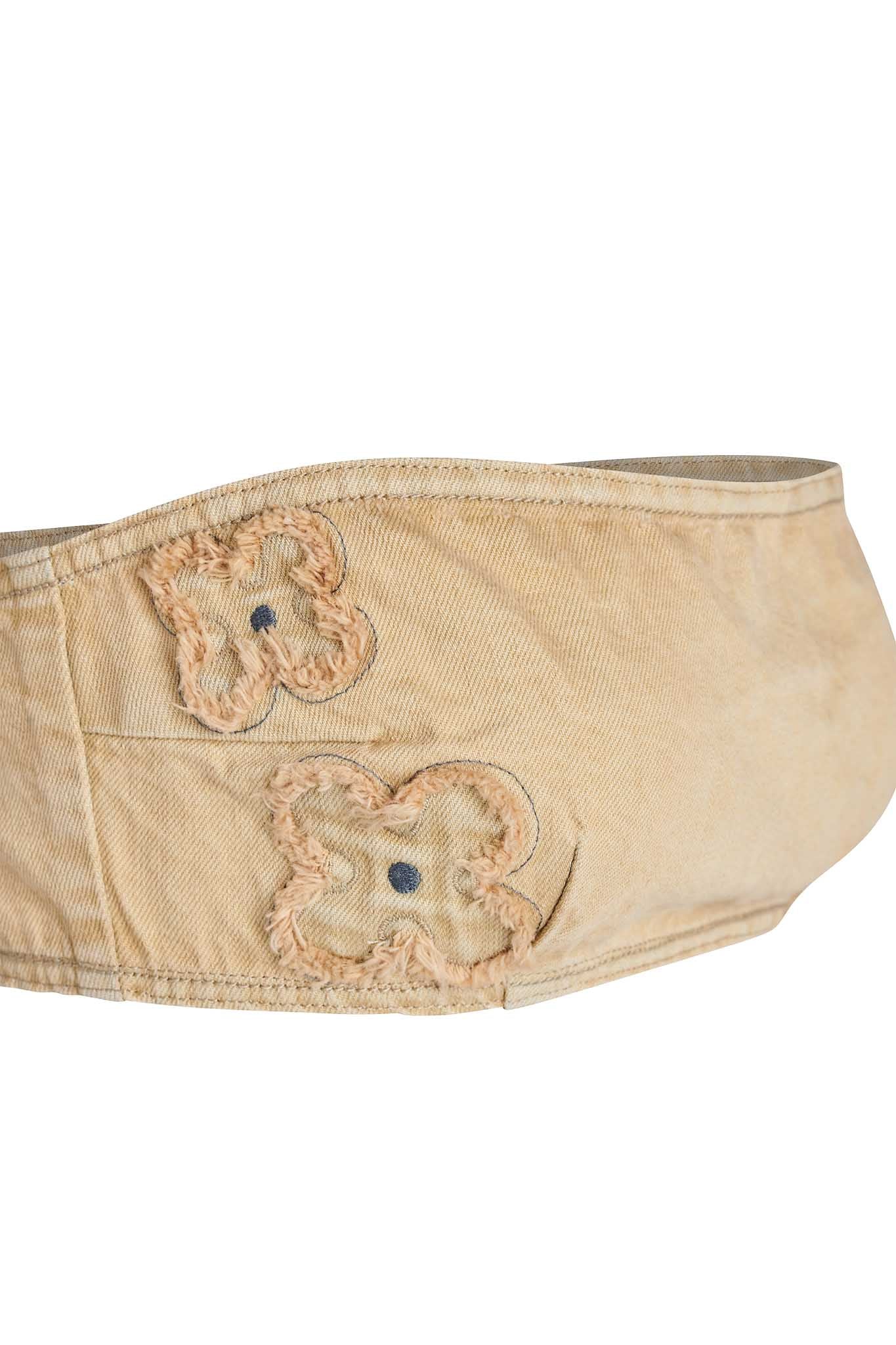 A vintage-inspired, beige bandeau crafted from tan denim with frayed embroidery, showcasing a few faint floral patterns accented by blue center dots. The fabric exhibits signs of wear and discoloration, evoking a sense of heavy use or age. This distinctive piece looks like it could be part of a belt or waistband and seamlessly complements the I.AM.GIA collection. 

Product Name: FAUNA BANDEAU - BEIGE : TAN DENIM
Brand Name: I.AM.GIA