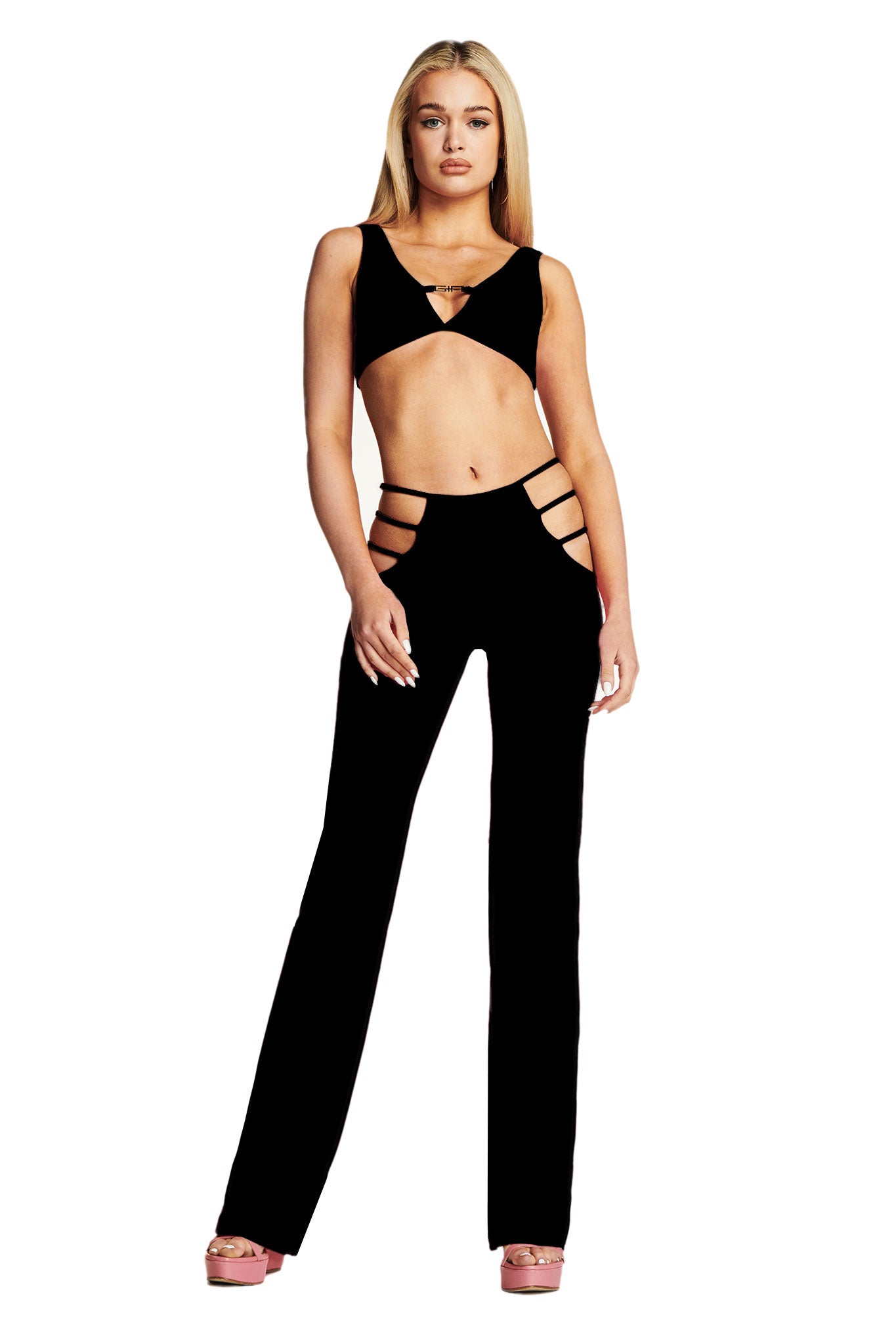 A woman with long blonde hair is standing and wearing the black I.AM.GIA LUCID TOP and matching black pants with side cutouts. She is posed confidently with one arm by her side and the other hand on her hip. She is also wearing pink platform heels.