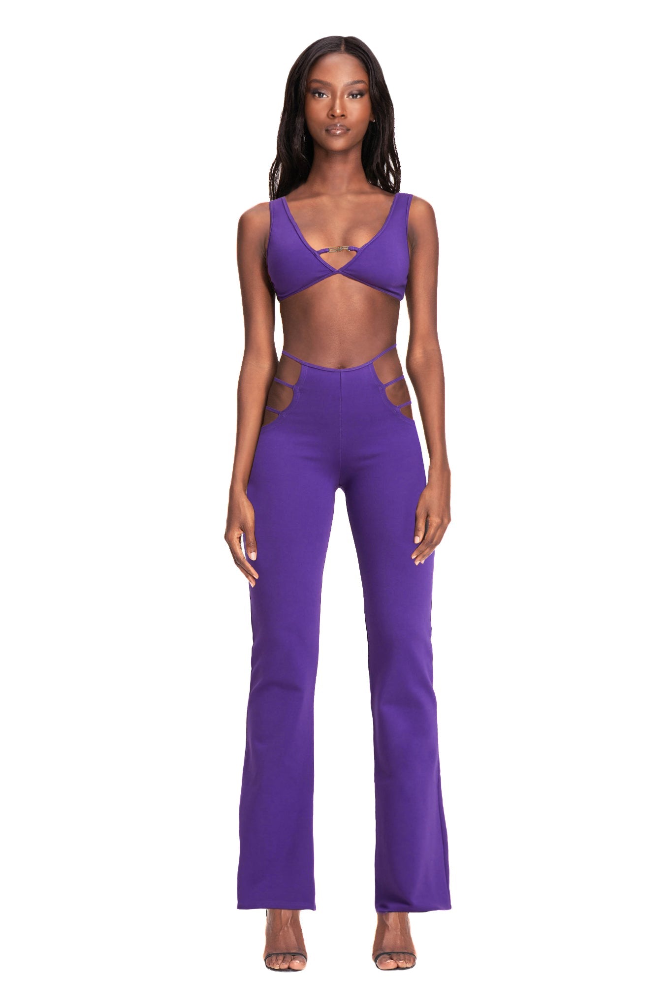 A woman stands against a white background, showcasing the I.AM.GIA LUCID PANT in purple. She wears a matching crop top with a crisscross design at the front and high-waisted flared pants featuring cut-out details on the sides. Her long, straight hair cascades down her back, and she completes the look with high-heeled sandals.